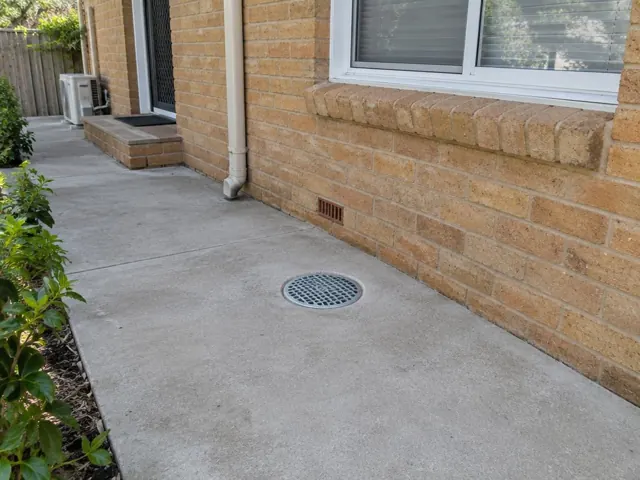 Round drain gully in a concrete patio beside a brick house wall, with dry paving in Turramurra