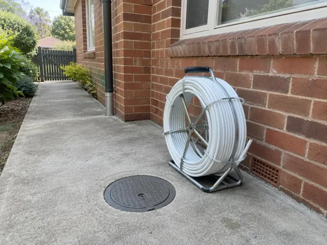 Capped access point in a concrete driveway beside a brick house with a pipe reel nearby in Wahroonga