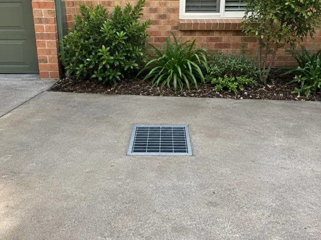 Square stormwater grate set in a concrete driveway beside a brick house and garden in Hornsby